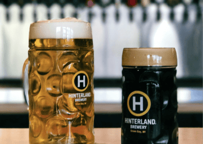 Two different sizes of Hinterland-branded glass steins full of beer sit on a bar.
